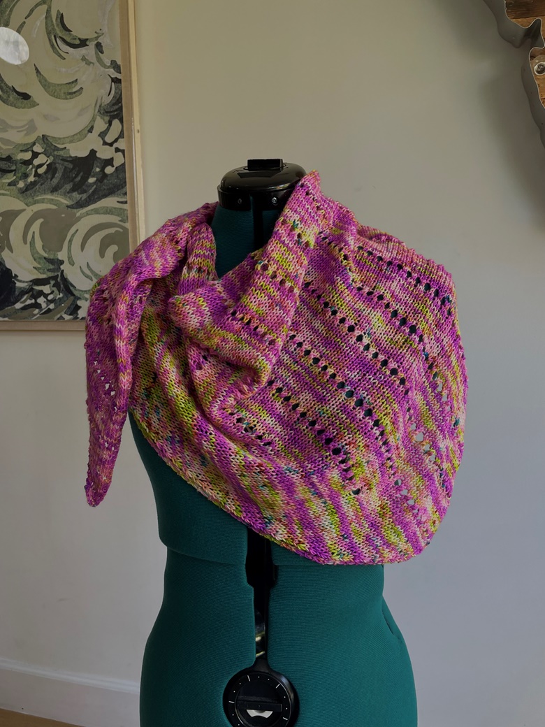 A bright knit shawl in pinks, yellows, and greens wrapped around the neck and shoulders of a green adjustable dress form