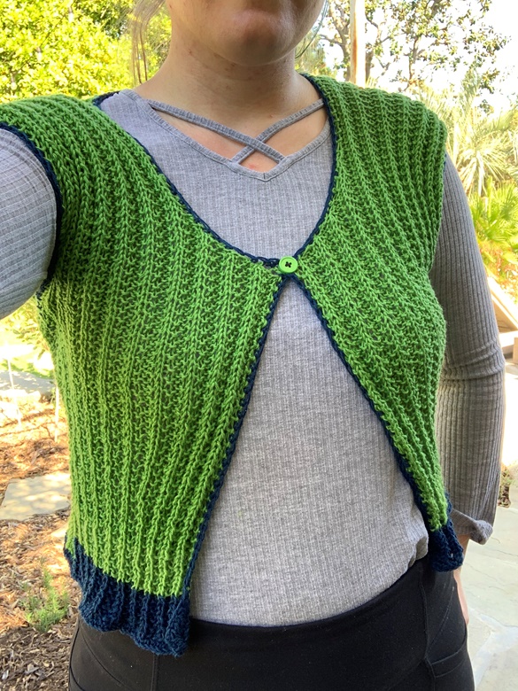 The torso of a woman wearing a green rib knit vest with a single green button at center neck, blue from hem, and blue crochet edging over a grey long sleeve shirt and black pants standing in a garden.