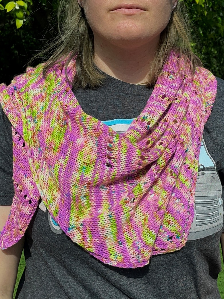 A close up of a knit shawl in pink, green, and yellow wrapped around the neck and shoulders of a white woman looking toward the camera
